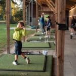 Jeugdige golfer maakt een swing op de driving range onder een overkapping, terwijl andere jeugdspelers en begeleiders trainen op de achtergrond.
