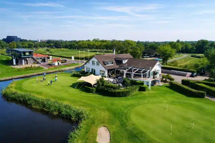 Luchtfoto van Golfbaan De Hoge Dijk met het clubhuis, een putting green met golfers, een terras en een waterpartij langs de fairway