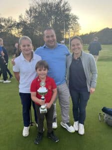 Jeugdige golfer met beker poseert samen met ouders en oudere speelster op de green na afloop van een golfwedstrijd bij zonsondergang.