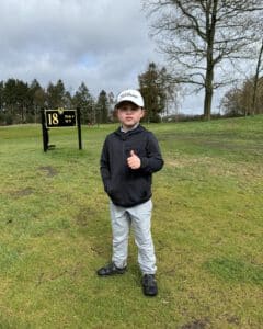 Jeugdgolfer staat op de fairway bij hole 18 (par 4) en geeft een duim omhoog, met baanbord en bomen op de achtergrond.