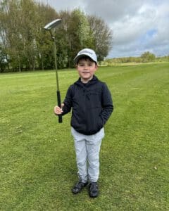 Jeugdgolfer staat op de putting green met een putter in de hand, gekleed in golfkleding, met fairway en bomen op de achtergrond.