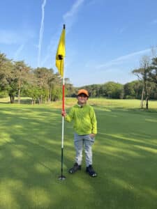 Jeugdgolfer staat glimlachend op de green en houdt de vlag vast bij de hole, met een zonnige golfbaan en bomen op de achtergrond.