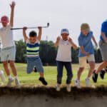 Groep jeugdige golfers springt juichend tegelijk in de lucht langs de rand van een bunker tijdens een vrolijke jeugdgolftraining op de golfbaan.