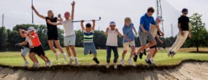 Groep jeugdige golfers springt juichend tegelijk in de lucht langs de rand van een bunker tijdens een vrolijke jeugdgolftraining op de golfbaan.